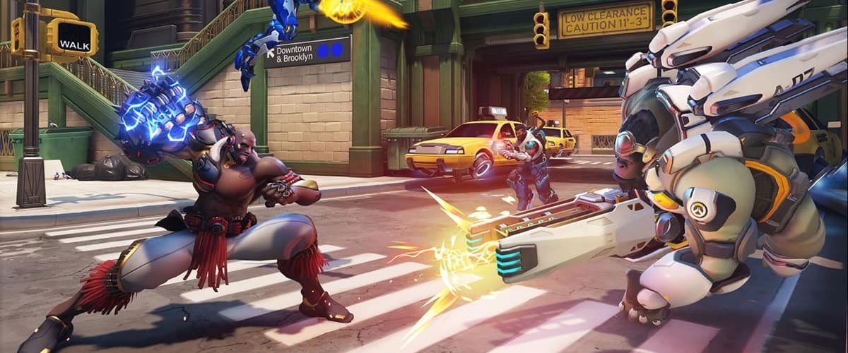 Un héros au poing mécanique fait face à un héros en armure blanche dans une rue new-yorkaise, une héroïne en armure bleue en vol au-dessus, dans Overwatch