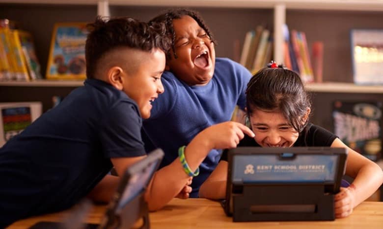 Trois élèves rient devant une tablette dans une salle de classe
