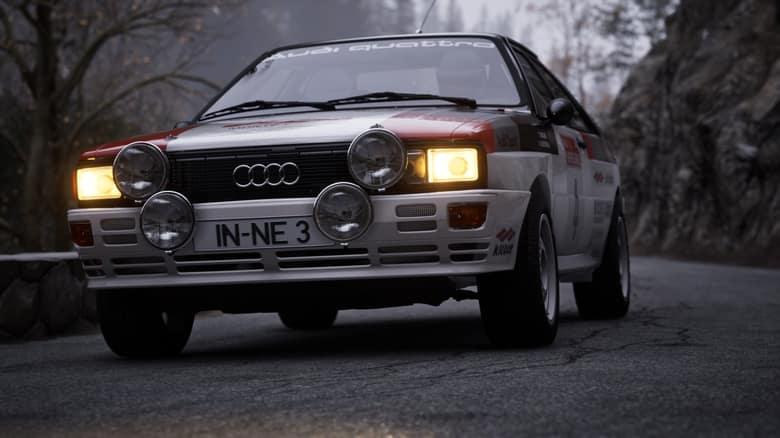 Une Audi Quattro blanche et rouge, phares allumés, sur une route de montagne